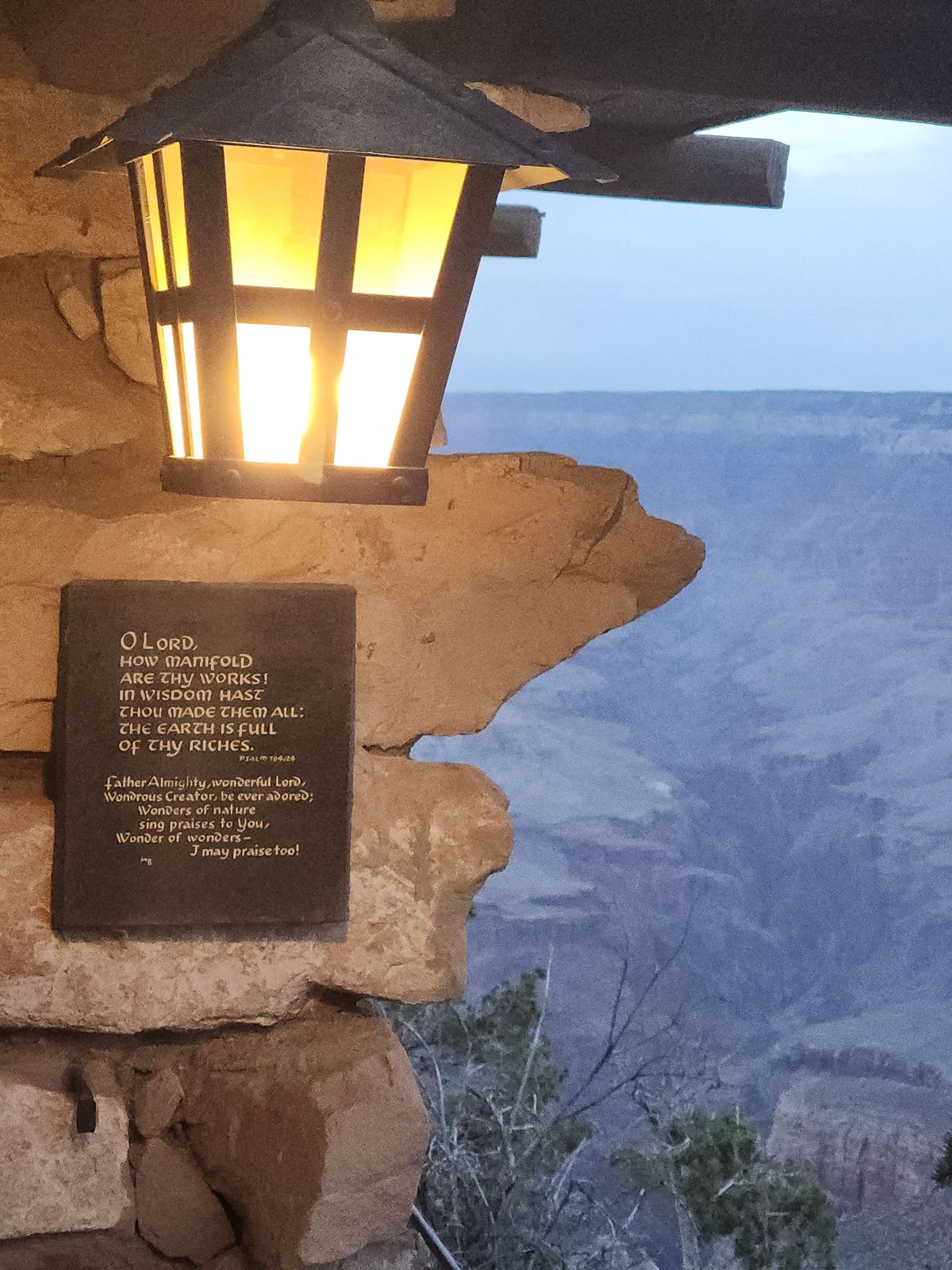 A lit lantern of iron and amber glass juts from a rough sandstone wall at dusk. Below it a bronze plaque carries an inscription. To the right the Grand Canyon falls away in layers of cold blue.